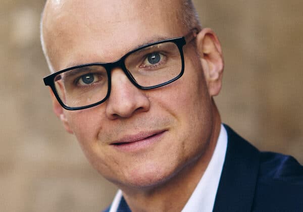 A man with dark-framed glasses smiles at the camera. He has blue eyes and light-toned skin. He is wearing a white shirt with a dark blue blazer.