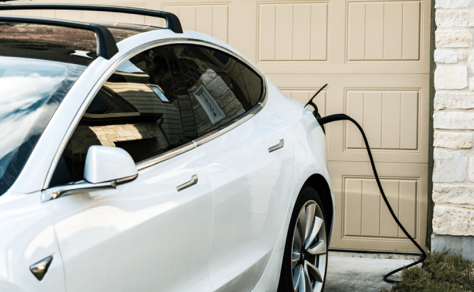 A white electric vehicle is parked in front of a house, charging from a residential charger.