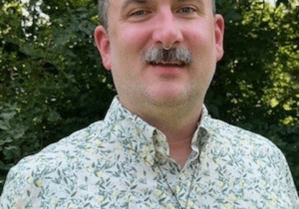 A man smiles at the camera. He has light-toned skin, salt and pepper hair, and a mustache. He is wearing a blue and green floral patterned shirt. Trees are in the background.