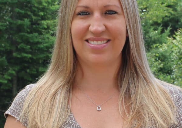 A woman with light-toned skin and straight blonde hair past her shoulders, smiles at the camera. She is wearing a short sleeve patterned shirt. Green trees fill the background.