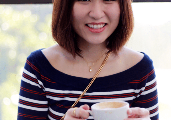 A woman smiles directly into the camera. She has a shoulder length brunette bob and is wearing a striped sweater. She is holding a cappuccino cup.