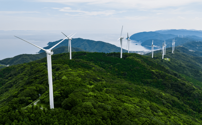 Wind turbines stretch across the ridge of a forested mountain range. In the distance is a large body of water.