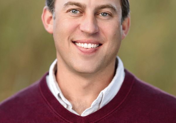 A man with short blonde hair smiles at the camera. He has light-toned skin and is wearing a maroon sweater over a collared shirt.