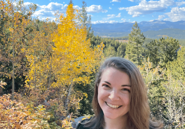 A woman with light-toned skin smiles at the camera. She has shoulder length blonde hair and is wearing a dark green sweater. A bright blue sky and changing fall leaves on a mountainside are behind her.