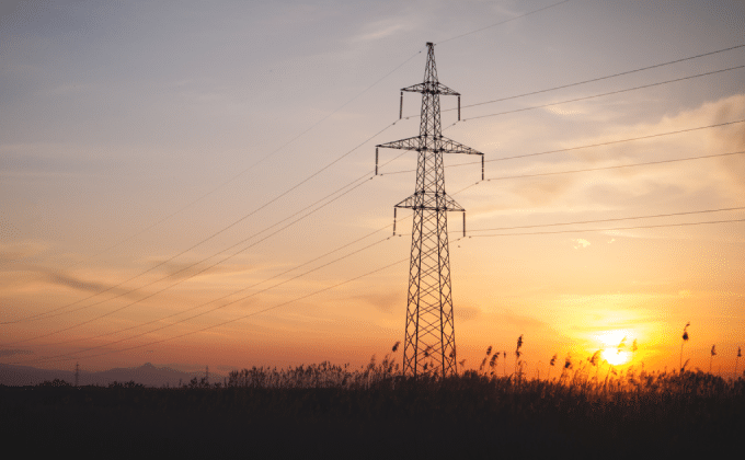 Transmission lines span across a field of wheat with an orange sunset in the background.