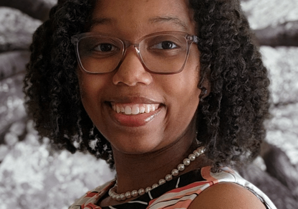 A woman with medium-dark skin smiles at the camera. She has black hair just above her shoulders and is wearing glasses and a pearl necklace.